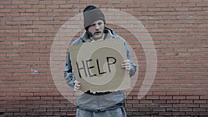 Homeless holds piece of cardboard with inscription Help. Refugee is at brick wall looking for job. Male tramp in dirty