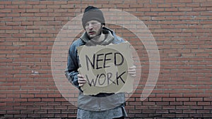 Homeless holds piece of cardboard with inscription Need Work. Refugee is at brick wall looking for job. Male tramp in