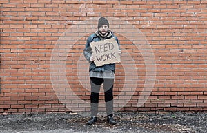 Homeless holds piece of cardboard with inscription Need Work. Refugee is at brick wall looking for job. Male tramp in dirty