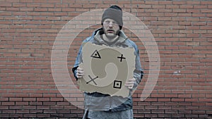 Homeless holds piece of cardboard with markers for planar tracking. Refugee is at brick wall looking for job. Male tramp