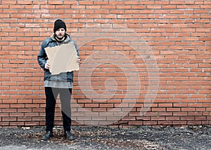 Homeless holds piece of cardboard. Refugee is at brick wall looking for job. Male tramp in dirty clothes. Below poverty line.