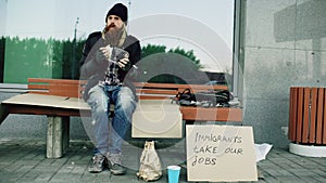 Homeless and jobless european man with cardboard sign eat sandwich on bench at city street because of immigrants crisis