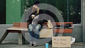 Homeless and jobless european man with cardboard sign eat sandwich on bench at city street because of immigrants crisis