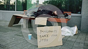 Homeless and jobless european man with cardboard sign sleep on bench at city street because of immigrants crisis in