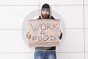 A homeless man with a beard and in a hood stands on the street with a sign about the request for work for food. Outdoor.