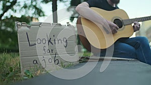 Homeless man with belongings on the street playing guitar, looking for a job