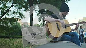 Homeless man with belongings on the street playing guitar, looking for a job