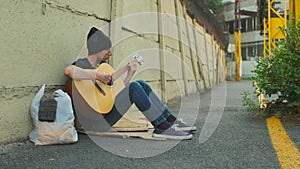 Homeless man with belongings on the street sitting by the fence playing guitar