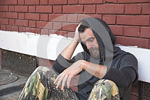 Homeless man drug and alcohol addict sitting alone and depressed on the street leaning against a red brick building wall