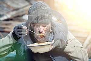 A homeless man eats soup from a plate near the ruins, helping poor and hungry people during the epidemic, sun glare on the image