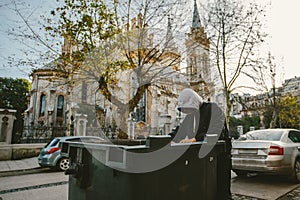 Homeless man looking into trash box in search for food on church background. Homelessness problem concept