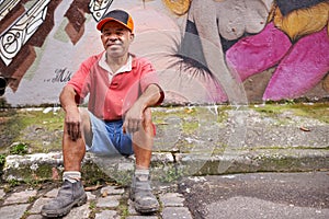 Homeless, man and poverty in street, mature male person and graffiti art on wall in urban area. Smile, sitting and