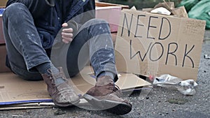 Homeless man, shivering from the cold, sits on a cardboard box among the garbage. Next to him is a handwritten poster