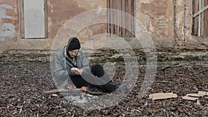 Homeless man sits on ground and pushes pieces of cardboard into fire to keep warm from cold. Tramp in dirty clothes and