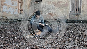 Homeless man sits on ground and pushes pieces of cardboard into fire to keep warm from cold. Tramp in dirty clothes and