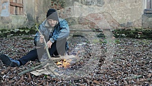 Homeless man sits on ground and uses stick to keep fire burning to keep warm from cold. Tramp in dirty clothes and hat