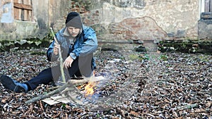 Homeless man sits on ground and uses stick to keep fire burning to keep warm from cold. Tramp in dirty clothes and hat cap. Below