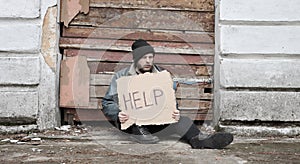 Homeless man sits and holds piece of cardboard with inscription Help. Refugee looking for job. Male tramp in dirty clothes, hat