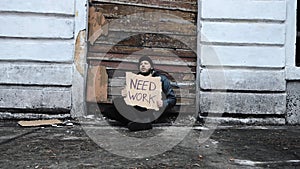 Homeless man sits and holds piece of cardboard with inscription Need Work. Refugee looking for job. Immigrant. Below poverty line