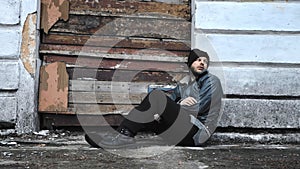 Homeless man sits outside an abandoned building. Consequences of drunkenness alcoholism. Tramp wearing in dirty clothes and hat