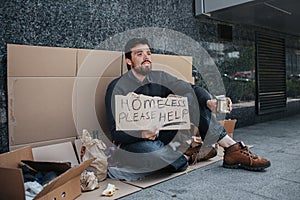 Homeless man is sitting on cardboard and and holding a sign that says homeless please help. He is begging money. Guy is