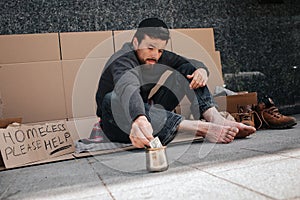 Homeless man is sitting on the cardboard outside and reaching metal cup with his hand. He is reaching a dollar. There is