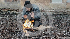Homeless man sitting and warming himself by the fire from cold. Tramp in dirty clothes and hat cap. Below poverty line