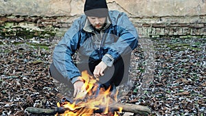 Homeless man sitting and warming himself by the fire from cold. Tramp in dirty clothes and hat cap. Below poverty line. Illegal