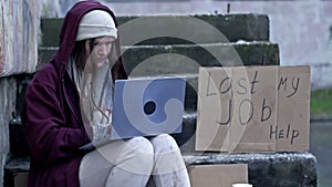 Homeless woman shivering in the cold with a laptop on her lap sits on cardboard amongst the rubbish. Next to her is a