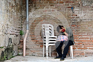 A homeless woman sleeping on a monoblock chair at the side of the brick wall in an old church yard