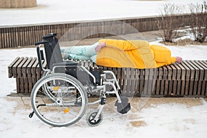 Homeless woman sleeping on a park bench next to a wheelchair.