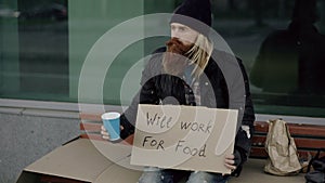 Homeless young man beg for money shaking cup to pay attention people walking near beggar at the city sidewalk