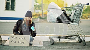 Homeless young man counting how much money he got holding paper cup in the hands and sitting near shopping cart at the