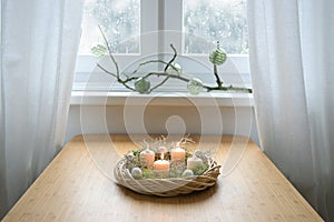Homemade advent wreath with Christmas balls, moss and white candles, the second one is lit, on a table near the window on a snowy
