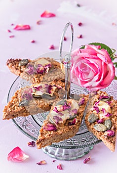 Homemade apple muesli bars with pumpkin seeds and rose petals on a light green glass cake stand on a delicate pink background
