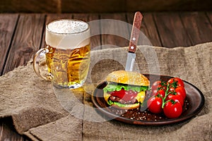 Homemade beef burger and fresh vegetables on Clay dish with glass of beer on rustic wooden table.