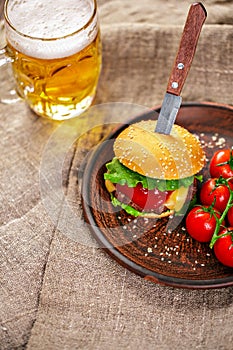 Homemade beef burger and fresh vegetables on Clay dish with glass of beer on rustic wooden table.