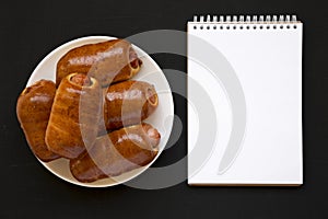 Homemade beef sausage kolache on a white plate, blank notepad on a black surface, top view. Flat lay, overhead, from above,