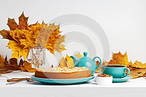 Homemade berry pie decorated with almond petals, cup of tea and yellow autumns leaves on white wooden table.