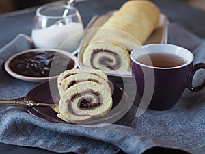 Homemade biscuit sponge roll with sweet plum jam and tea against a dark background. Natural homemade dessert, side view