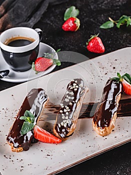 Homemade cake eclairs or profiteroles with custard, chocolate and strawberries on dark background served with cup of coffee.