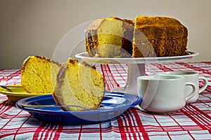 Homemade cake with yellow and blue dishes on the side and cups of coffee on the table with white and red checkered tablecloth. Hom