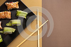 Homemade california and cucumber sushi rolls with sesame seeds on a black square plate on a brown beige background with wooden
