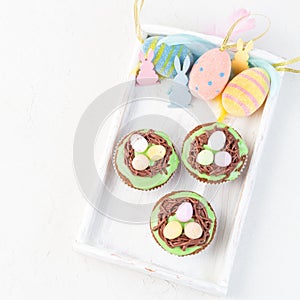 Homemade carrot cupcakes with cream cheese frosting and Easter decoration, on a white wooden tray, top view, square