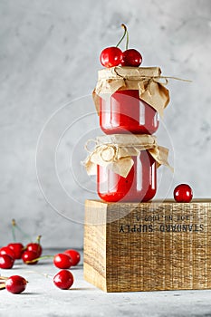 Homemade cherry jam in glass jar on the wooden box on the gray background. Two jars of cherry jam on wood with cherries in the