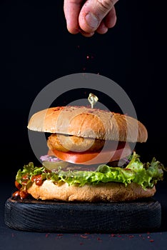 Homemade chicken burger on serving dark wooden board and man's fingers sprinkling red pepper