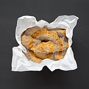 Homemade chicken fingers in paper box on a black surface, top view. Flat lay, overhead, from above. Close-up