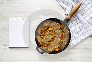 Homemade Chicken Tikka Masala in a cast iron pan, blank notepad on a white wooden background, top view. Flat lay, overhead, from