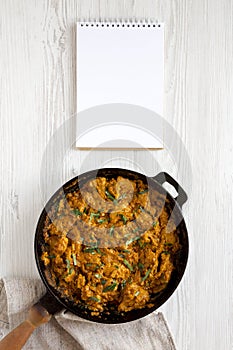 Homemade Chicken Tikka Masala in a cast iron pan, blank notepad on a white wooden background, top view. Flat lay, overhead, from