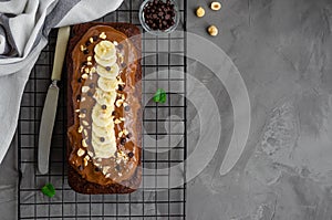 Homemade chocolate banana bread with chocolate cream, banana slices and nuts on top on a dark concrete background.